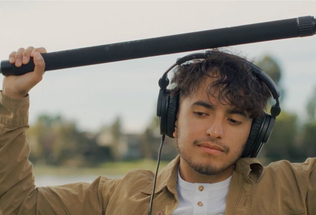 A young man wearing headphones holds a boom pole over his head.
