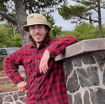 a young man in a red flannel leans casually against a wall