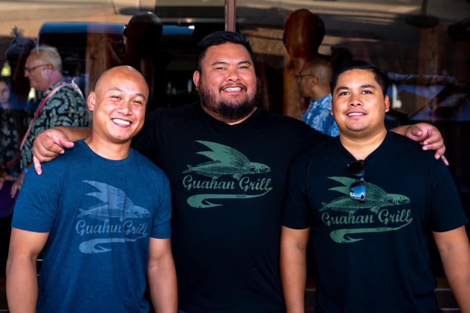 The owners of Guahan Grill stand next to each other, wearing merchandise from their restaurant