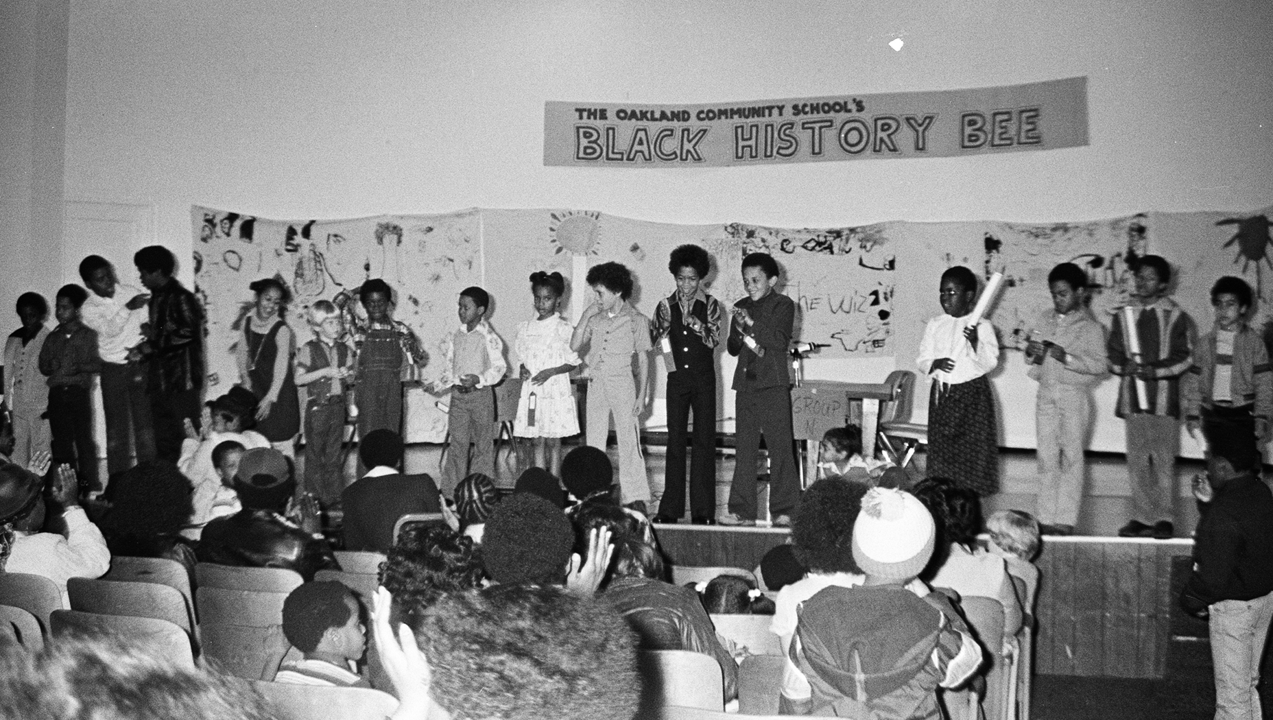 A black and white photo taken at the OCS at a Black History Bee. A group of students stand up on stage in front of an audience.