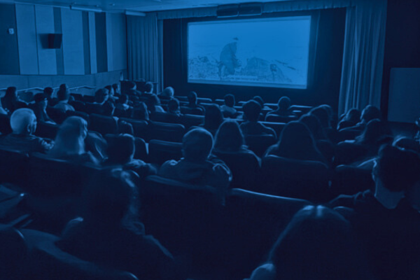 A dimly lit movie theater filled with a diverse audience watching a film. The screen displays a scene with a person in a landscape setting, creating an engaged and focused atmosphere.