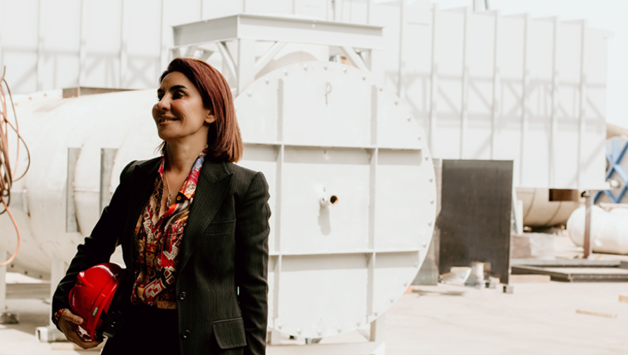 Anoosheh Oskouian is in an industrial plant. She is holding a red hard hat and wearing a colorful blouse and black blazer. She is facing the left side of the frame and smiling