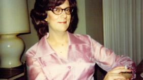 Transgender person seated with dark hair, pink satin dress 1970s