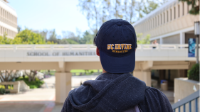 A transfer student with a UC Irvine Humanities baseball cap looking at the School of Humanities bridge.