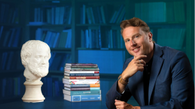 Duncan Pritchard is siting in a library at a table with a pile of books and a philosophers bust. The background has a blue overlay. 