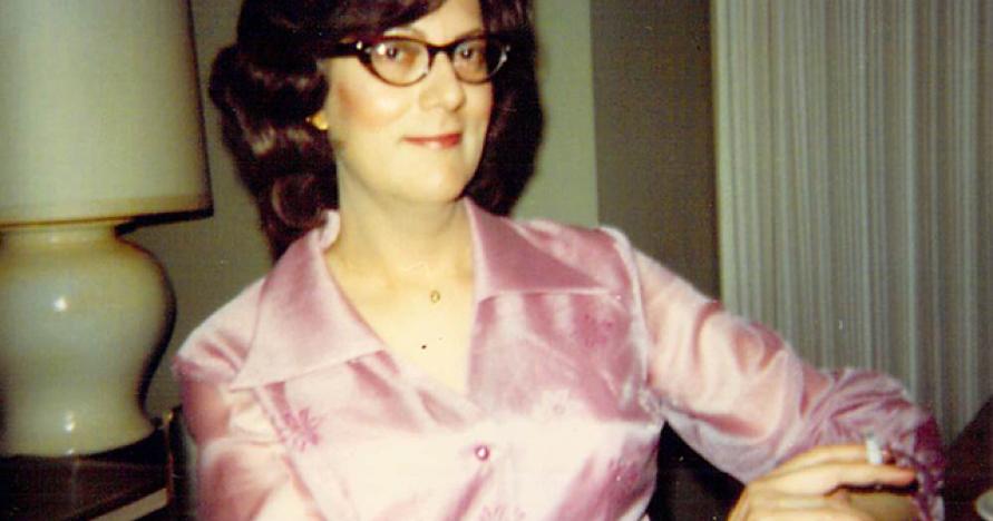 Transgender person seated with dark hair, pink satin dress 1970s