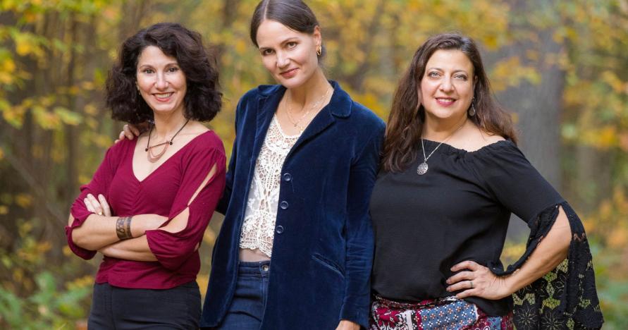 The three women who make up the band Zulal stand next to eachother smiling in front of trees.