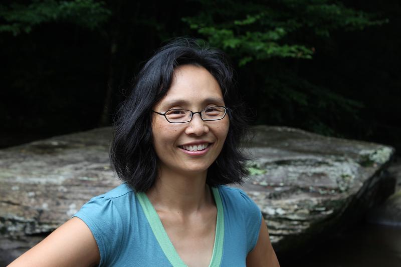 Head and shoulders of woman with dark hair and teal short sleeve shirt standing next to stream