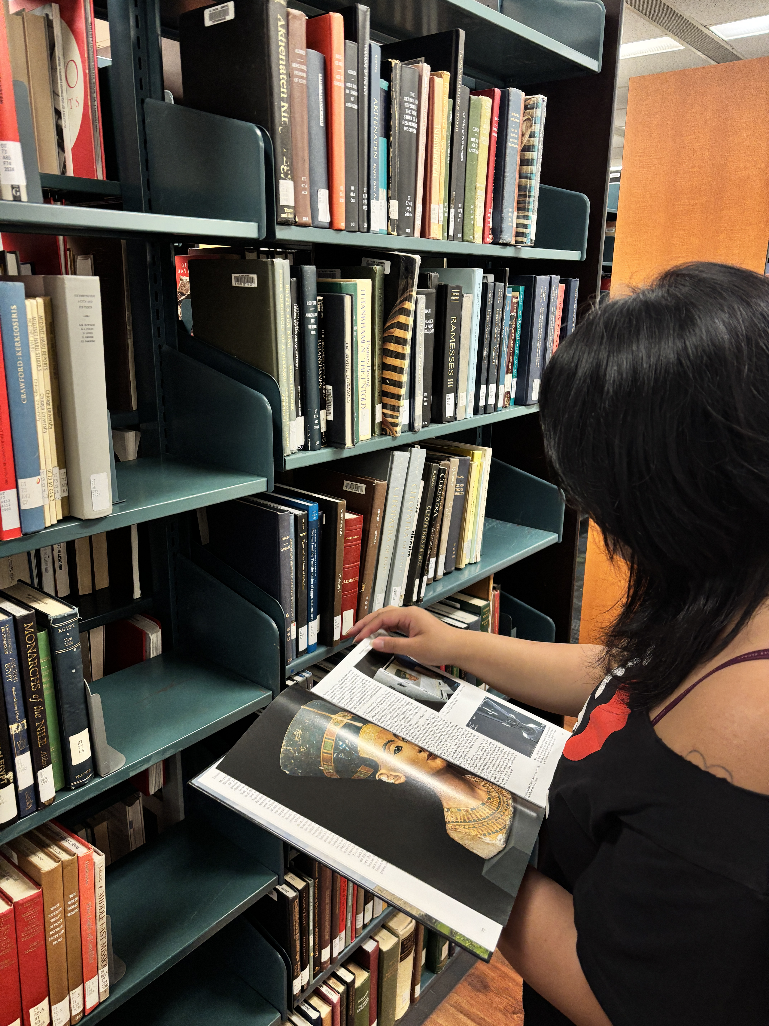 Sofia is reading a book and standing next to a book shelf.