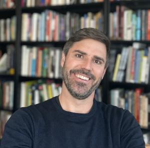 Headshot of smiling man wearing black sweater in front of bookshelf
