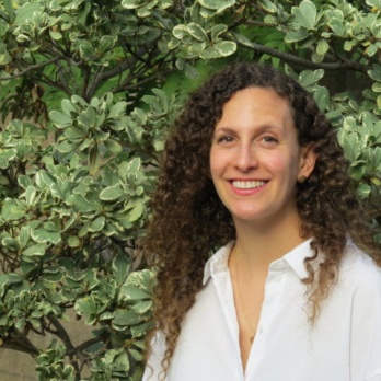 woman with long curly brown hair wearing white shirt in front of green bush