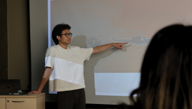 An action shot of UTeach student Brian Nguyen in the classroom talking about his powerpoint slide.